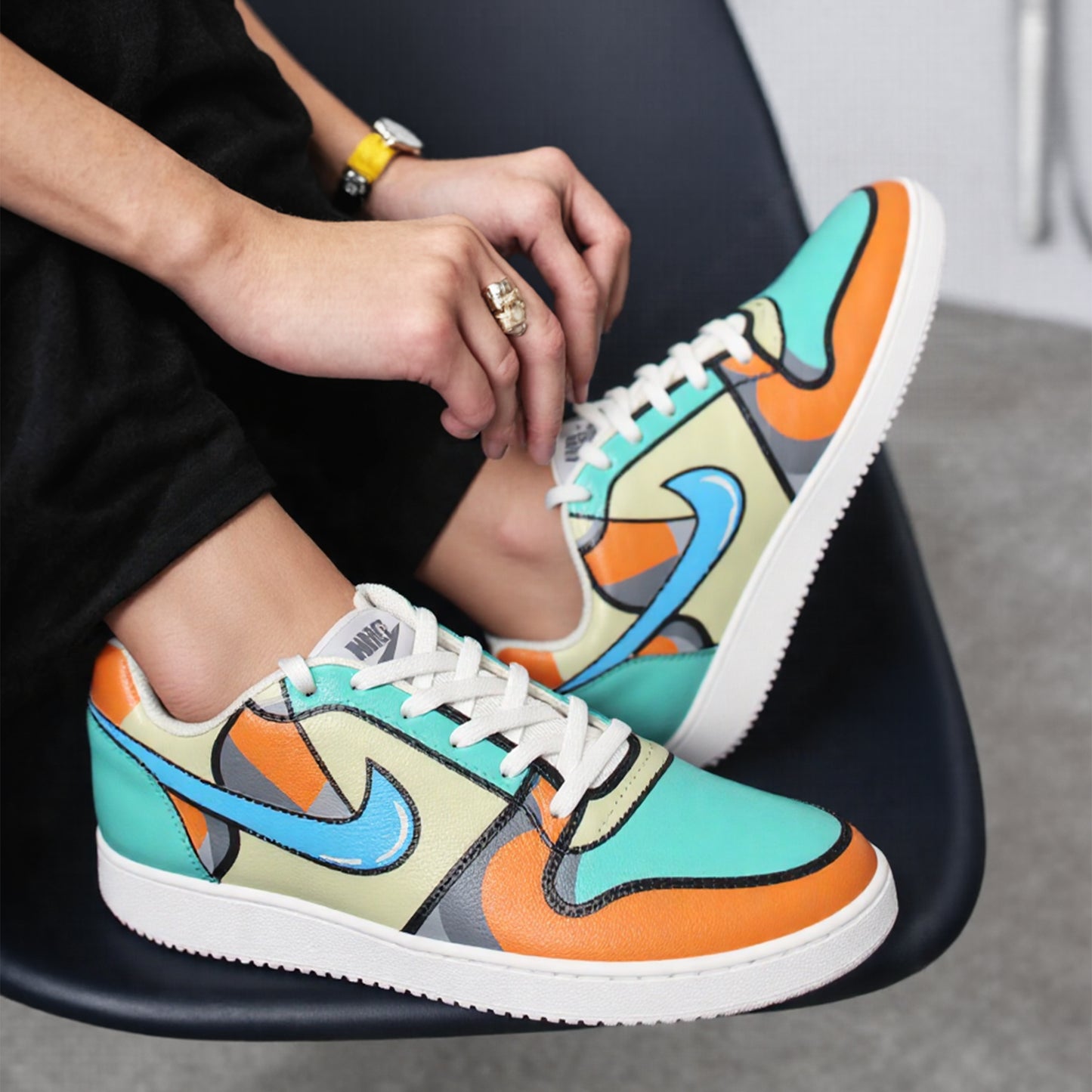Hand-painted Nike Colourful sneaker with a unique geometric design in orange, grey, beige, and blue colours, worn by a person sitting on a black chair.