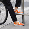 Person wearing hand-pinted orange and red sneakers with a yellow swoosh logo, standing next to a bicycle on a sidewalk.